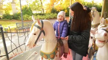 Uzun siyah saçları ve küçük kızı sürme ile mutlu anne Carousel ve birlikte gülüyor. Bebek kız esmer kadın yanındaki bir dolambaçlı bir beyaz atı üzerinde oturan mor ceket.