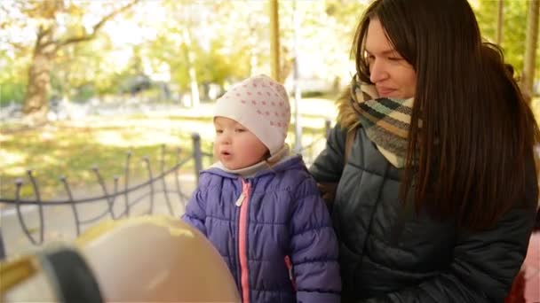 Усміхаючись, мати і малюк їзда на карусель в Осінній Парк протягом доби. Красива жінка і мила дівчина, які мають задоволення разом на відкритому повітрі сидить на білому коні в кільцевої — стокове відео
