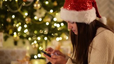 Noel Baba şapkası siyah Smartphone kullanarak ve arka plan Noel ağacına gülümseyen güzel kadın. Güzel kız onun Gadget Xmas Cap yazarak iletide. Arka plan bulanık Noel ışıkları