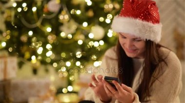 Noel Baba şapkası esmer cep telefonunu yazarak üzerinde gülümseyen arka plan ve Noel ağacı. Gadget'ı kullanarak Xmas şapkalı kız. Ladin bulanık ışıkları.