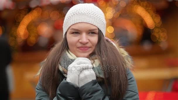 Ritratto di una ragazza ridente che indossa un cappello bianco e guanti grigi sullo sfondo delle luci di Natale, mani riscaldanti donna in inverno. Buon Natale e Felice Anno Nuovo — Video Stock