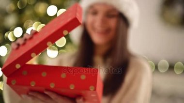 Noel Baba şapkası hayretler içinde kırmızı bir kutu içinde sürpriz ile güzel kadın. Sevimli gülümseyen kız Noel hediyesi Xmas ağacı arka planda, Closeup açma.