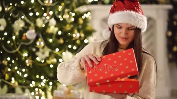 Glücklich erstaunt Frau mit Weihnachtsmütze, hält Geschenk in ihren Händen, öffnet es und lächelt. hübsche Brünette mit langen dunklen Haaren und Weihnachtsmannmütze zum Öffnen der Weihnachtsgeschenkschachtel neben dem Weihnachtsbaum — Stockvideo