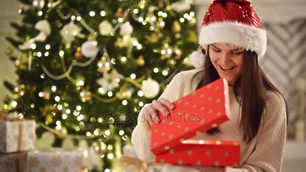 Joven, Feliz y Emocional Chica en Santa Sombrero Abriendo el Regalo Mágico de Navidad. Pretty Lady in Red Cap abre caja con regalo al lado del árbol de Navidad en casa decorada y ríe — Vídeo de stock