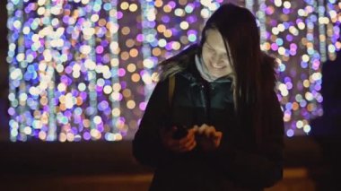 Smartphone Noel ışıkları arka plan üzerinde gece şehirde güzel bir kadınla kış koyu ceket. Uzun saçlı esmer gülümseyen sokakta duran Gadget'ı kullanır. Bulanık Xmas Garland, Dusk.