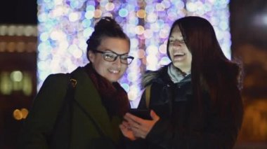 Cep telefonunuzun ve Noel ile bir gece sokakta kız gülümseyerek dekorasyon ışıklar. İki esmer kız Gadget ekrana bakarak gülüyor. Bulanık Xmas Garland.