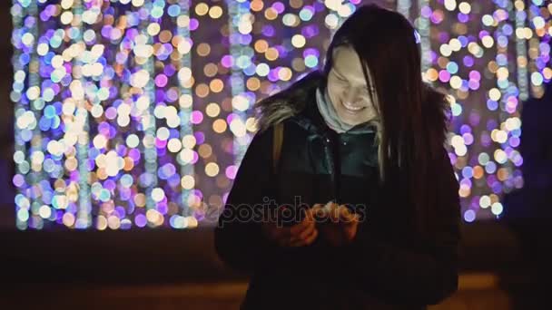 Bella donna in inverno scuro cappotto con smartphone nella città di notte su sfondo luci di Natale. Bruna sorridente con capelli lunghi utilizza gadget in piedi per strada. Ghirlanda di Natale offuscata, Crepuscolo . — Video Stock