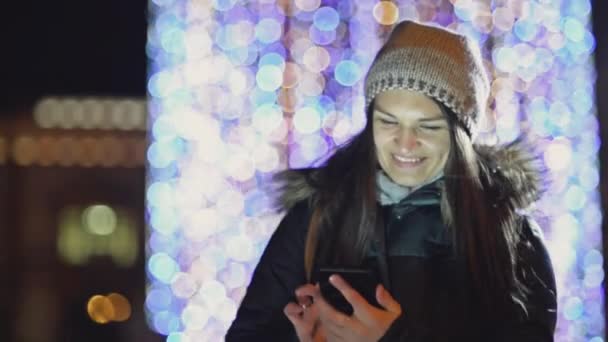 Młoda Pani w kapelusz stojąc na ulicy i przy użyciu telefonu komórkowego. Uśmiechnięte dziewczyny z gadżetu na zewnątrz na tle światła Bożego Narodzenia. Niewyraźne Boże Narodzenie girlanda, Zmierzch. — Wideo stockowe