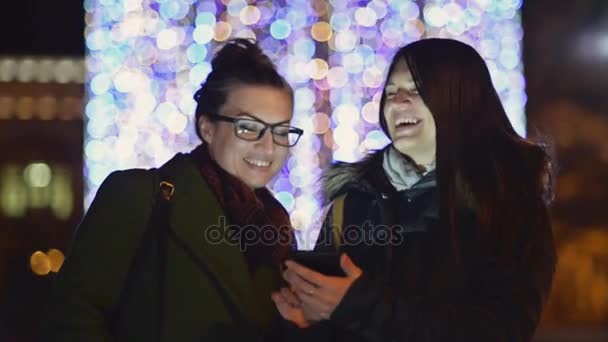 Sœurs souriantes utilisant le téléphone portable sur une rue de nuit avec décorations lumières de Noël. Deux filles brunes riant en regardant l'écran Gadget. Guirlande de Noël floue . — Video