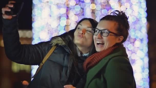 Dos amigas tomando un selfie al aire libre en el fondo de las luces de Navidad. Mujeres jóvenes que se fotografían afuera usando Smartphone. Guirnalda de Navidad borrosa . — Vídeos de Stock