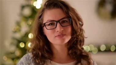 Mutlu genç kadın, gözlük takıyor, gösterilen dişlek gülümseme Noel ağacı arka kamera karşı yukarı kapatın. Kıvırcık uzun saçlı güzel Bayan portresi. Bulanık Xmas Lights.