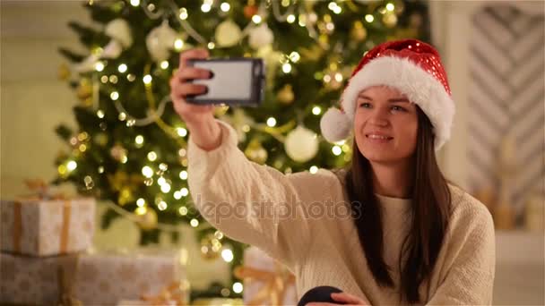 クリスマスのお祝いの中にスマート フォンで Selfie をしているサンタ帽子の若い幸せな女。かわいい女の子のクリスマス ツリーの背景に自分自身を撮影. — ストック動画