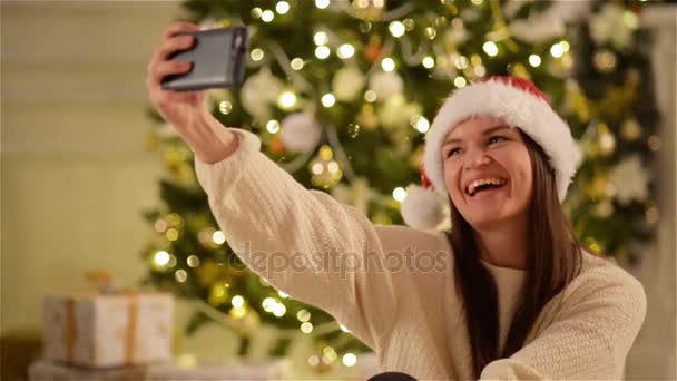 Χριστούγεννα πορτρέτο του χαμογελαστό γυναίκα φοράει καπέλο Santa και καθιστώντας Selfie στο κινητό τηλέφωνο. Χαριτωμένο κυρία με μακριά σκούρα μαλλιά, φωτογραφίζοντας τον εαυτό της σχετικά Gadget. Xmas δέντρο φόντο. Θολά φώτα. — Αρχείο Βίντεο