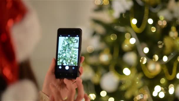 Pani w Santa Hat robi migawkę choinki przez smartfona. Piękne Dekoracje świąteczne świerk — Wideo stockowe