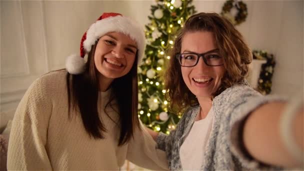 2 つのクリスマス ツリーの背景に Selfie を作る魅力的な女性の笑みを浮かべてします。眼鏡をかけて、巻き毛を持つ一人の女の子別のサンタ帽子. — ストック動画