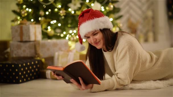 Πορτραίτο closeup χαμογελαστό κορίτσι που κρατά ένα ανοιχτό βιβλίο στα χέρια δίπλα στο χριστουγεννιάτικο δέντρο και τα κουτιά με τα δώρα. Ευτυχισμένη γυναίκα κατάπληκτος με το καπέλο Santa με μακριά μαλλιά βάζοντας στο πάτωμα και να απολαύσετε την ανάγνωση. — Αρχείο Βίντεο