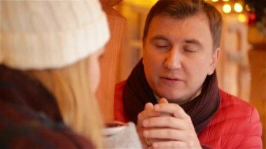 Kırmızı ceket ve sıcak içki ve kadın ile bir şey hakkında komik konuşuyor bordo eşarp gülümseyen adam. Mutlu kaç Noel çarşı sırasında bir sokak Cafe kahve içme sıcak giysiler içinde.