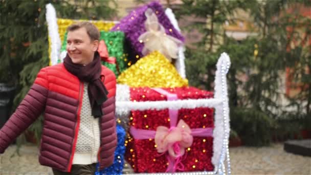 Ein Mann und eine Frau halten Händchen und gehen am Weihnachtsmann-Schlitten mit bunt verpackten Geschenken auf dem Weihnachtsmarkt vorbei. ein verliebtes Paar beim gemeinsamen Spazierengehen im Freien. — Stockvideo