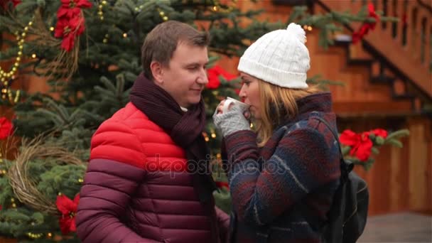 Hombre guapo y mujer guapa bebiendo café en el mercado de Navidad de la tradición. Esposa y esposo se miran con amor y ternura tomando bebidas calientes al aire libre en el fondo del árbol de Navidad . — Vídeo de stock