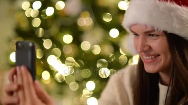 Mutlu kadın Noel Baba şapkası Xmas ağacı arka plan üzerinde Noel kutlamaları sırasında Selfie, yapıyor. Kendini Smartphone kullanarak çekim kız gülümseyerek closeup portresi.