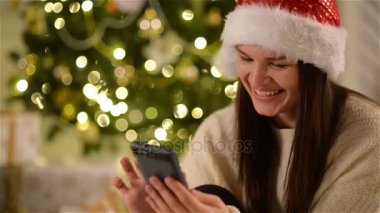 Gülümseyen Santa Noel ağacın yanında oturan Gadget'ı kullanarak şapkalı kadın hayran. Uzun siyah saçlı bir ekran cep telefonu, ev dekore Xmas dokunmadan neşeli kız.