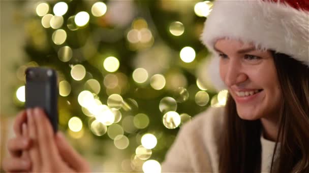 Gelukkige vrouw in KERSTMUTS Selfie, tijdens de viering van Kerstmis op Xmas boom achtergrond doen. Closeup portret van glimlachen meisje dat zichzelf met behulp van Smartphone fotograferen. — Stockvideo