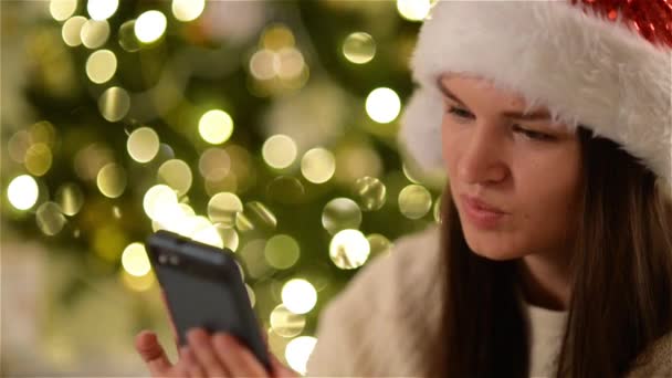 Morena usando Santa Sombrero está buscando algo en el Smartphone, Tener una sonrisa encantadora en el fondo del árbol de Navidad. Emocional chica sonriendo y sosteniendo el gadget en la mano durante la celebración de Navidad . — Vídeo de stock