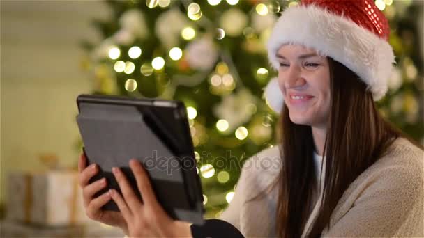 Linda senhora no chapéu de Santa chama alguém usando a câmera de vídeo no tablet durante a celebração de Natal no fundo da árvore de Natal . — Vídeo de Stock