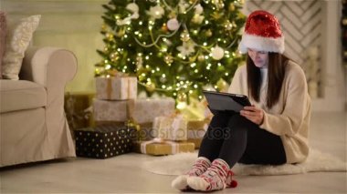 Noel Baba şapkası Tablet, Noel ağacının yanındaki katta halı üzerinde oturan kullanma esmer kadın. Noel kutlamaları sırasında elinde Gadget ile gülümseyen kız.