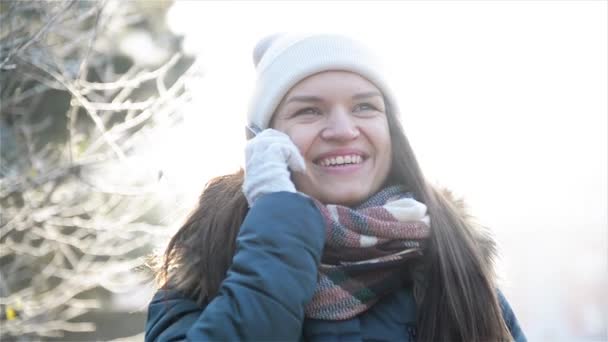 Mooi meisje in een witte hoed en geruite sjaal gesprek met behulp van mobiele telefoon op winterlandschap. Jonge lachende vrouw praten met iemand door Smartphone in zonnige ochtend Outdoors. — Stockvideo