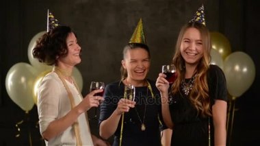 Grup genç bayanlar içme şampanya ve şarap altın kapaklar ve güzel elbiseler giymiş bir partide. Gülümseyen kadın kız hava balonu siyah zemin üzerine ile doğum günü kutluyor.