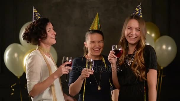Gruppen av unga damer på en fest dricka Champagne och vin bär gyllene Caps och vackra klänningar. Leende kvinna firar födelsedag med sina väninnor på svart bakgrund med Air ballonger. — Stockvideo