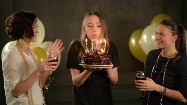 Tres hermosas mujeres celebran y aplauden, una de ellas sosteniendo pastel de chocolate, otras dos copas con vino. Cumpleaños Chica sopla las velas. Fondo negro con globos de aire . — Vídeos de Stock