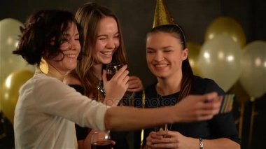 Güzel kadınlar ellerindeki Smartphone kullanarak kendilerini fotoğraf çekme gözlüklü. Gülümseyen kız beyaz ve altın hava balonları ile siyah arka plan üzerine bir partide bir fotoğraf çekmek için dijital Gadget'ı kullanma.