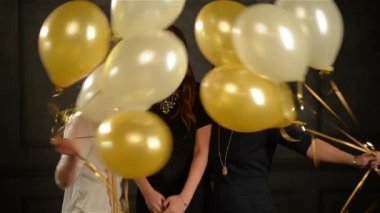 Sürpriz kadınlar arasında eğlence sırasında parti balonları gizleme heyecanlı. Siyah arka plan üzerine stüdyoda üç gülümseyen kız closeup portresi.