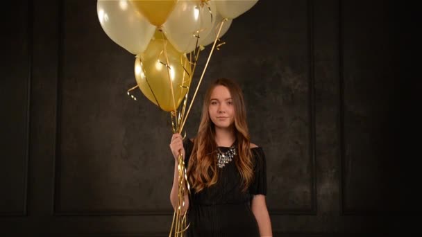 Party Girl z powietrza balony w jej ręce stojący wśród konfetti złotej i srebrnej. Uśmiechnięta młoda kobieta z długimi włosami świętować coś, czarne tło. — Wideo stockowe