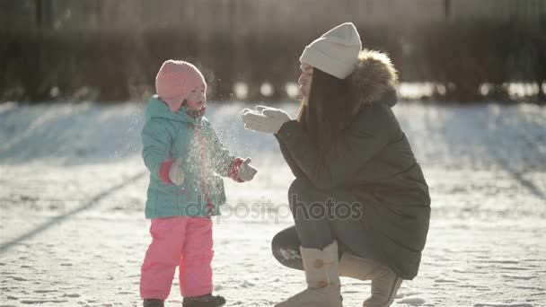 かわいい小さな女の子を手から雪を吹き飛ばし、彼女の母。冬は屋外の日当たりの良い寒さで朝散歩中に幸せな家族. — ストック動画