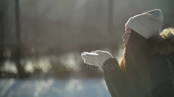 Úžasná žena v profilu je foukání na sněhové vločky z její ruce stojící v zasněžené zimní Park v dopoledních hodinách. Hravá dívka se těší chladné slunečné počasí venku. — Stock video