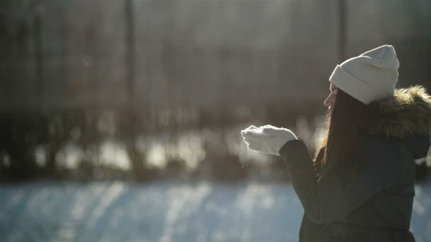 Garota brincalhona desfrutando de tempo ensolarado frio fora. Mulher incrível em perfil está soprando em um flocos de neve de suas mãos de pé no parque de inverno nevado na parte da manhã . — Vídeo de Stock