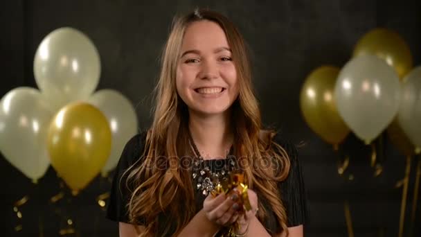 Primer plano Retrato de chica bonita que está soplando en Confetti. Mujer joven con pelo largo y collar sopla en pequeños pedazos de papel dorado sobre fondo negro con globos de aire . — Vídeos de Stock