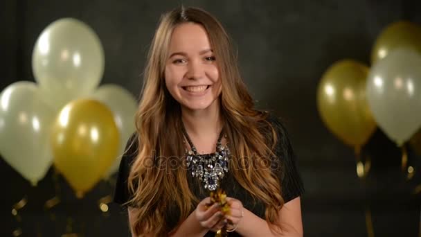 Joyeuse femme aux cheveux longs et collier souffle sur confettis sur fond noir avec des ballons à air. Portrait de mignonne fille de fête qui souffle sur de petites pièces de papier doré . — Video