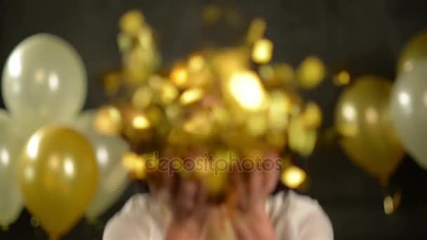 Retrato de mulher bonita com curly cabelo preto curto que está soprando em confetti dourado. Brincos brincalhões durante a celebração no estúdio sobre fundo com balões . — Vídeo de Stock