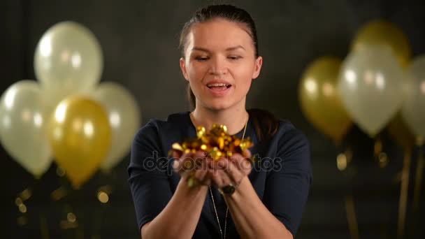 Femme élégante souffle sur Confetti qui tombe lentement vers le bas. Brunette pendant la célébration en studio sur fond noir avec des ballons à air doré et blanc . — Video