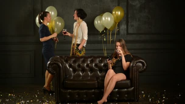 Chica triste en vestido corto y tacones altos está utilizando el teléfono inteligente a la fiesta sentada en el sofá junto a las mujeres que están hablando. Damas alegres discutiendo con otra porque ella las está ignorando . — Vídeos de Stock