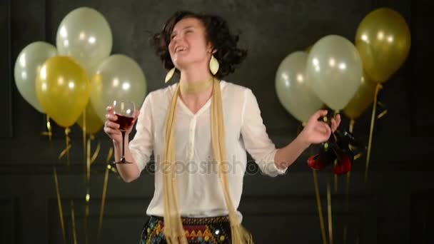 Joyous Party Girl Apreciando Dança entre Confetti em Fundo Preto com Balões. Morena encantadora com cabelo curto encaracolado está dançando segurando copo de vinho e sapatos de salto alto vermelhos nas mãos . — Vídeo de Stock