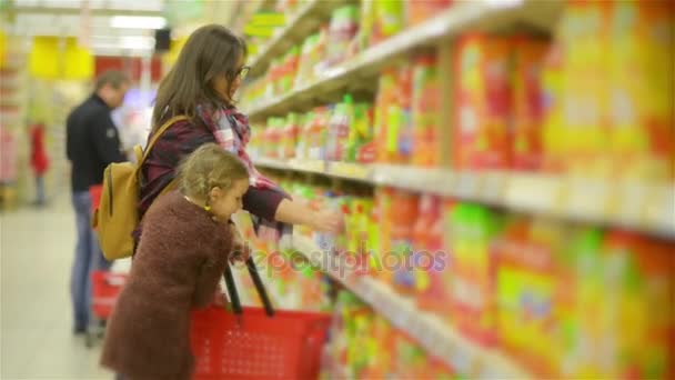 Junge Mutter und kleine Tochter beim Einkaufen im Supermarkt. Mutter und Kind putzen gemeinsam Produkte auf den roten Korb im Laden. — Stockvideo
