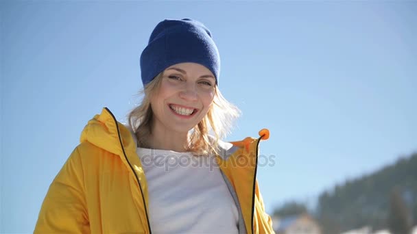 Gros plan Portrait de jolie blonde portant une veste jaune vif et un chapeau foncé à l'extérieur pendant la journée ensoleillée d'hiver. Une femme souriante profite du beau temps sur fond de ciel bleu . — Video