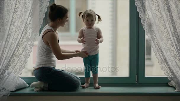 Side View of Loving Mother with Charming Smile and Cute Daughter with Two Ponytails hugging each other next to the Window. Маленькая девочка стоит на подоконнике, удивительная женщина обнимает ее . — стоковое видео