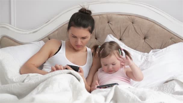 漂亮妈妈和她可爱的女儿，与两个马尾辫是使用小工具在一起躺在白色的床。美丽的黑发女郎在手智能手机的小女孩举行。. — 图库视频影像