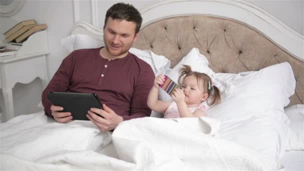 Padre guapo y su pequeña hija están acostados en la cama y el uso de dispositivos electrónicos. Niña con colas de caballo sostiene Smartphone, su padre está mostrando algo en la pantalla de la tableta . — Vídeo de stock